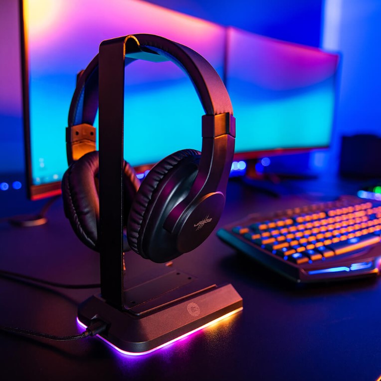 uRage gaming headset hängandes på ett stativ med RGB belysnings samt 2st datorskärmar i bakgrunden och ett tangentbord